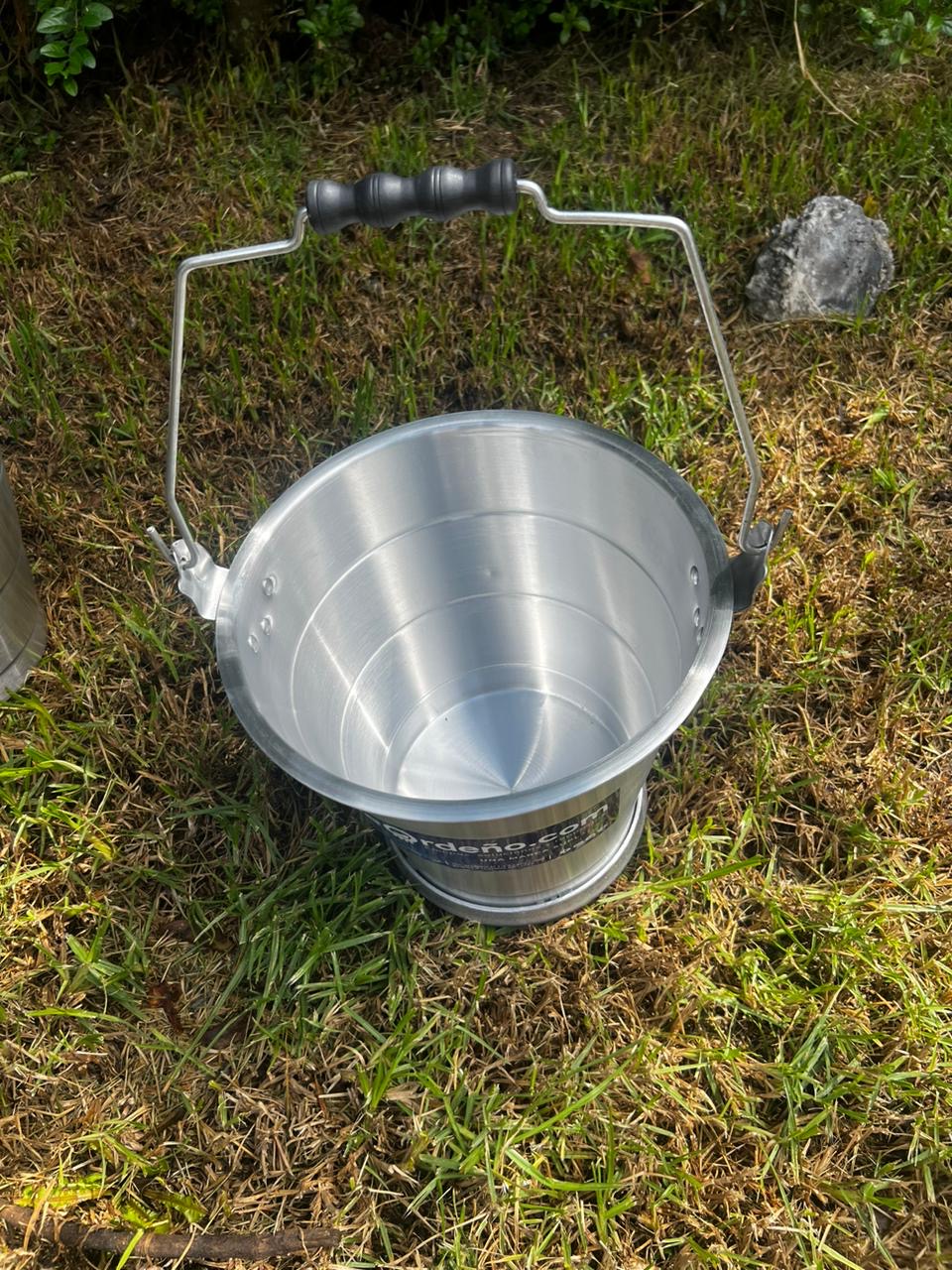 Balde lechero de aluminio apoyado en el suelo, mostrando el cuerpo y el asa